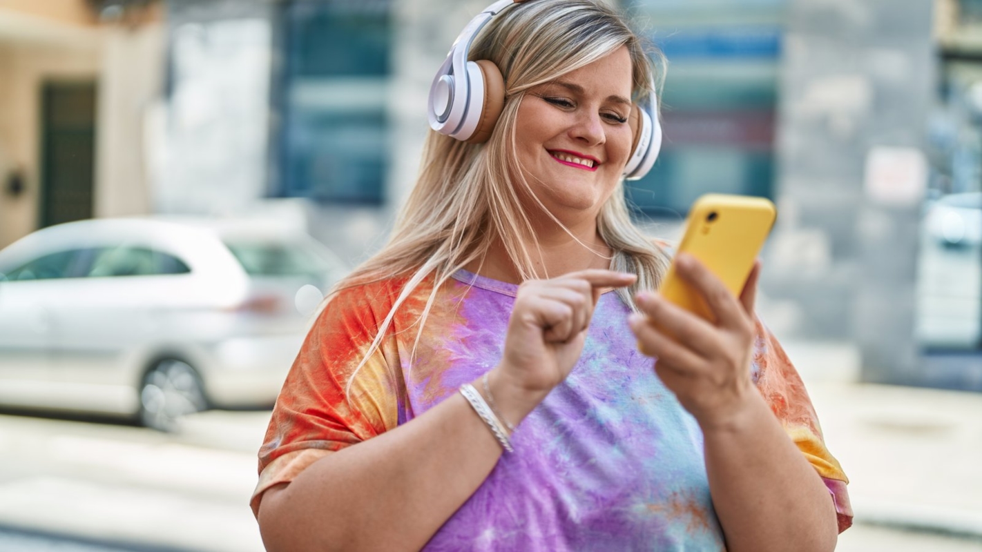 A woman smiling while listening to headphones and looking at her phone on a city street