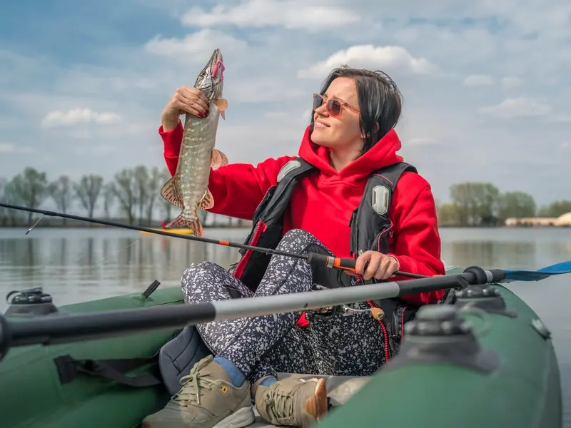A smiling woman in a red hoodie holding up a pike she caught from her inflatable kayak