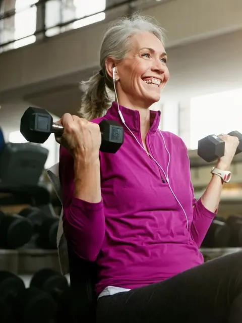 A smiling silver-haired woman lifting dumbbells in a bright gym