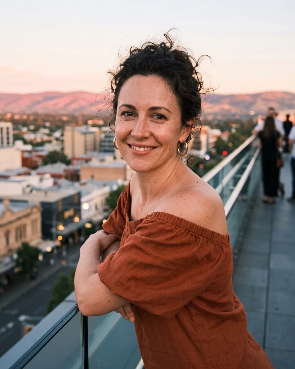 A woman with dark curly hair pinned up, gold hoops and an off-the-shoulder rust linen dress leans on a rooftop balustrade in Adelaide at sunset, hills glowing behind her (AI-generated image)