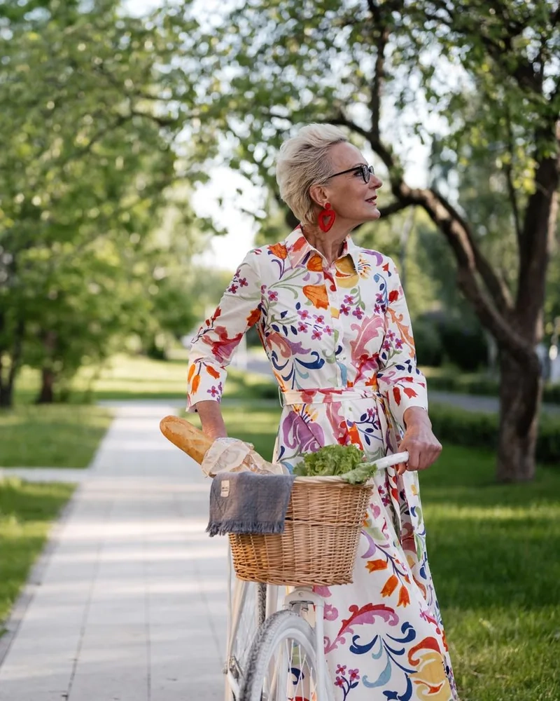 A woman in a colourful dress walking beside her bicycle, a basket of bread and greens on the handlebars, along a sunny park path