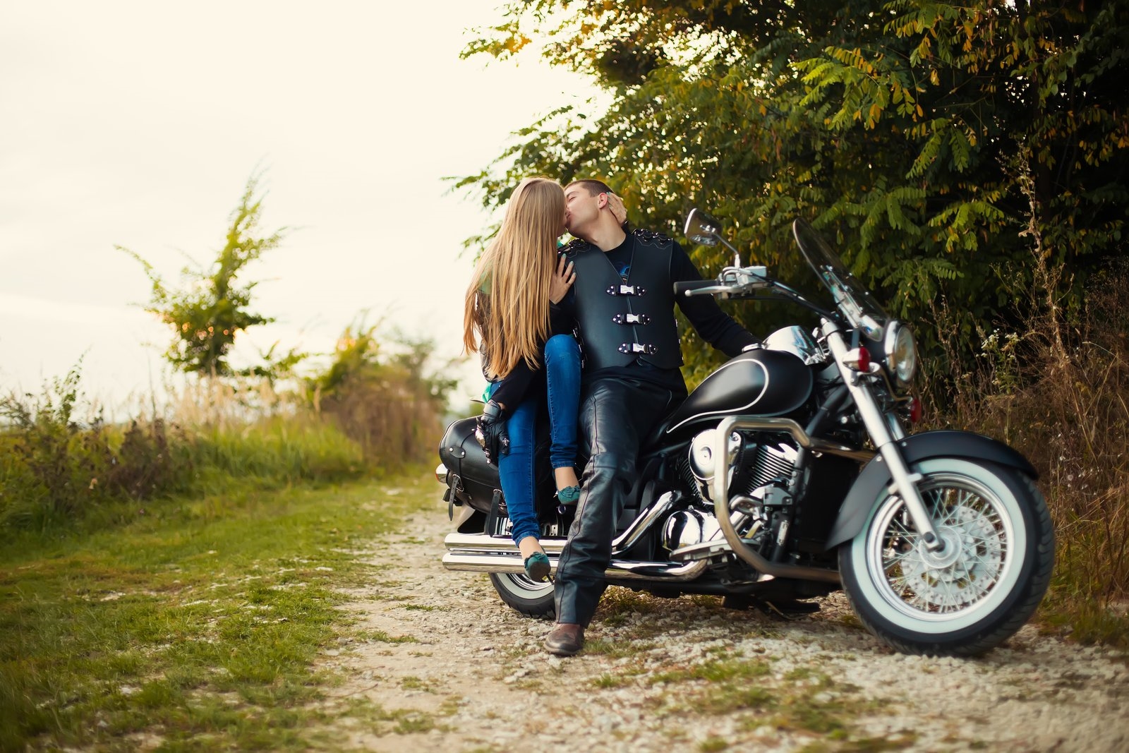 A couple kissing while leaning against a cruiser motorcycle on a tree-lined dirt path