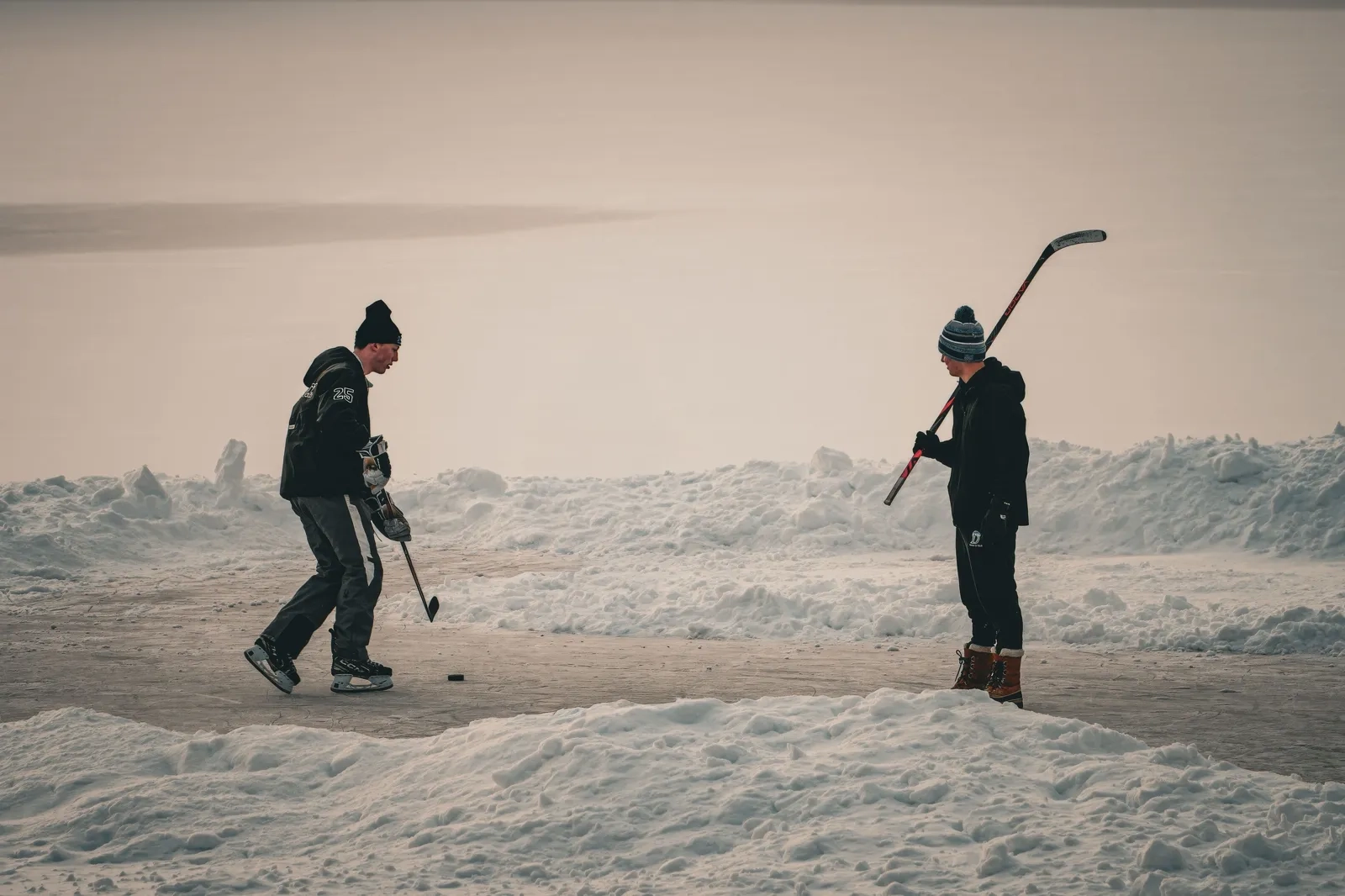 Two people playing hockey on a frozen, snow-dusted lake at dusk
