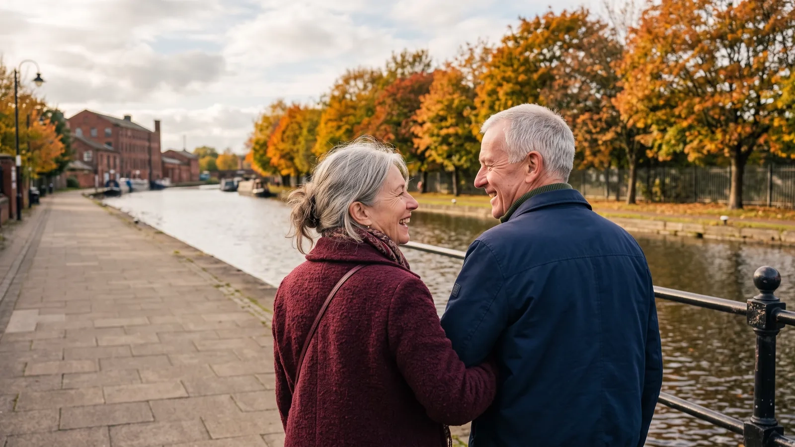 A couple in their late sixties walk arm in arm along a waterside path beside a canal lined with autumn trees and old red brick buildings (AI-generated image)