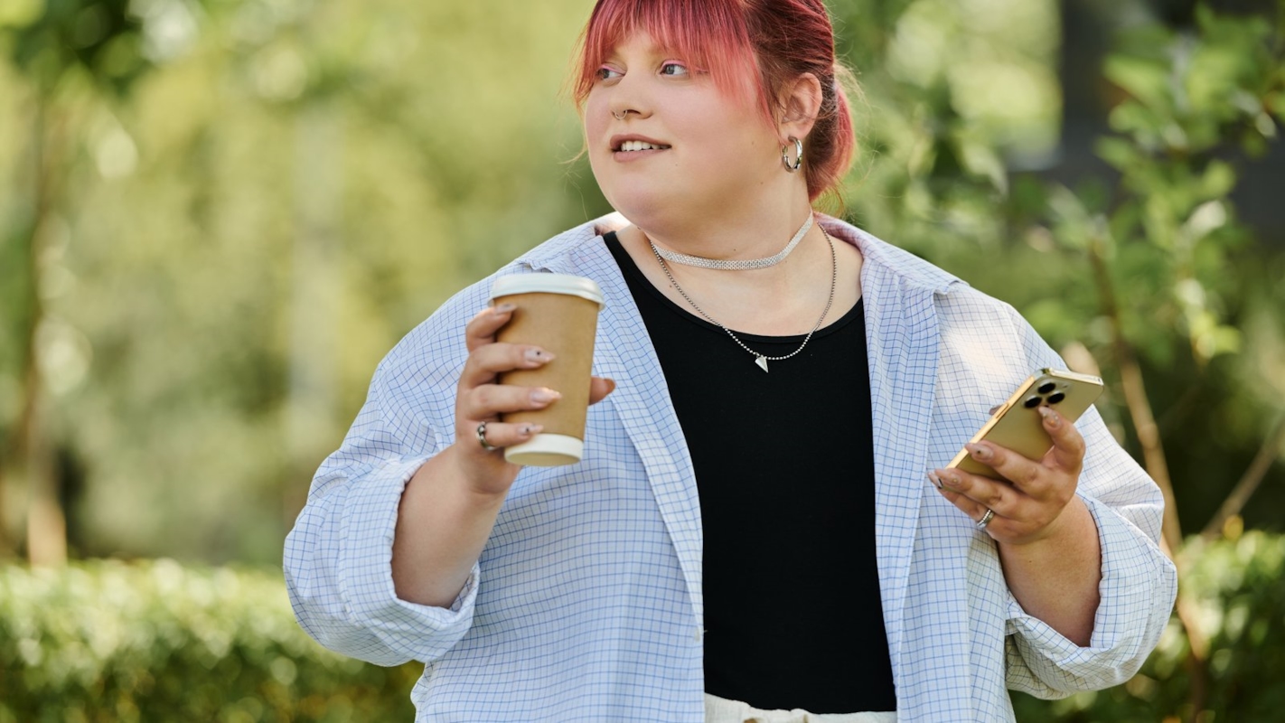 A woman smiling outdoors with a coffee and her phone