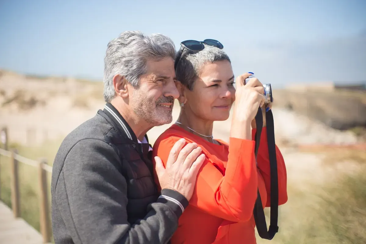 A smiling couple on a sunny boardwalk by the dunes, the woman taking a photo with a camera