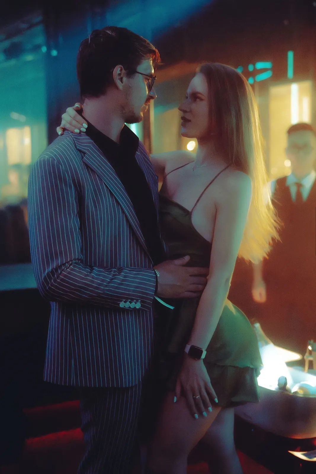 A man in a striped blazer and a woman in a dark dress standing close together in a softly lit bar, illustrative photo of models