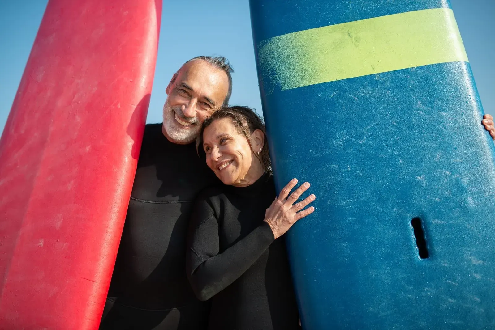 A couple in wetsuits cuddling between their surfboards on a sunny beach