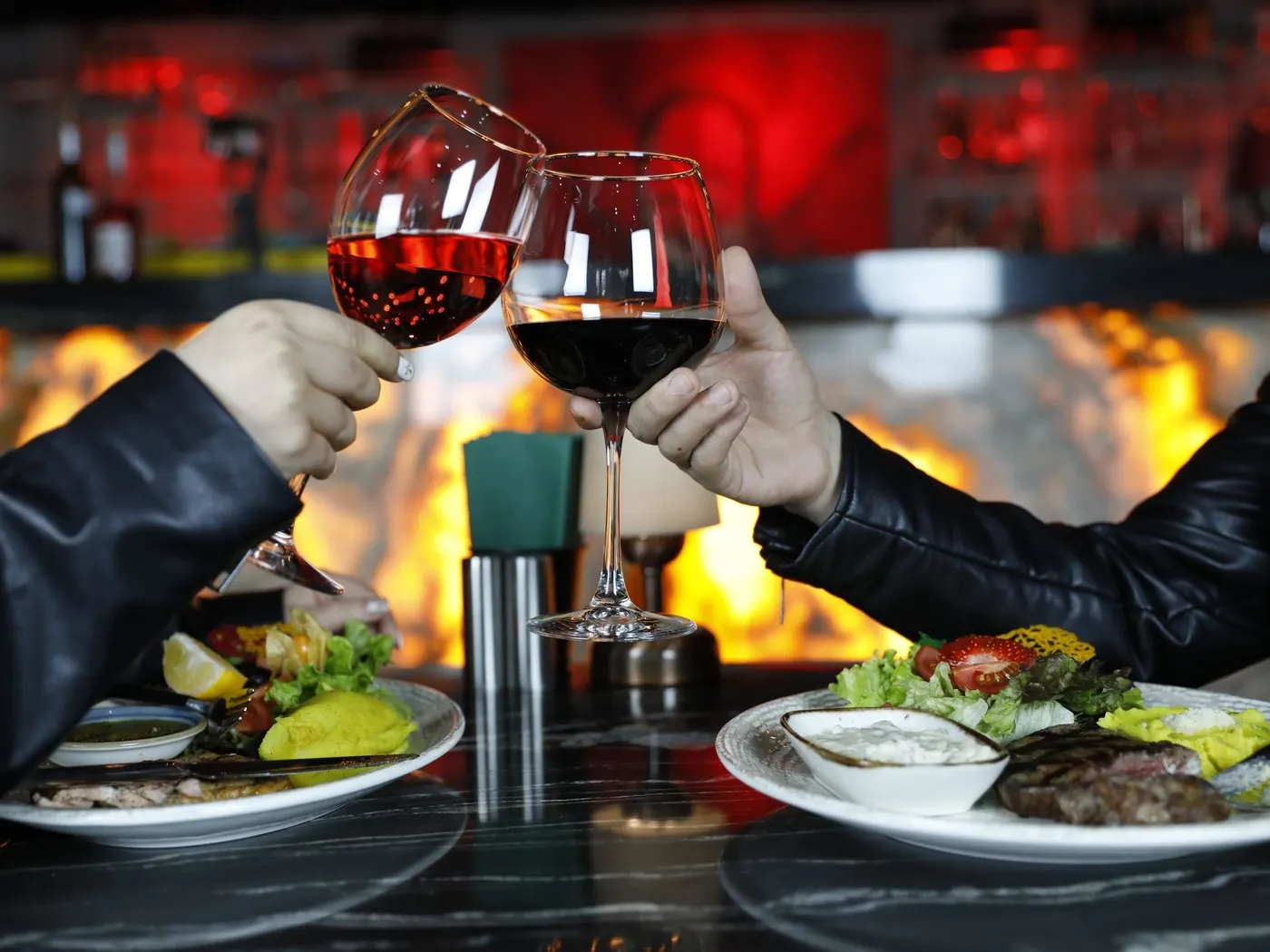 Two hands clinking wine glasses over a dinner table with a fire glowing behind