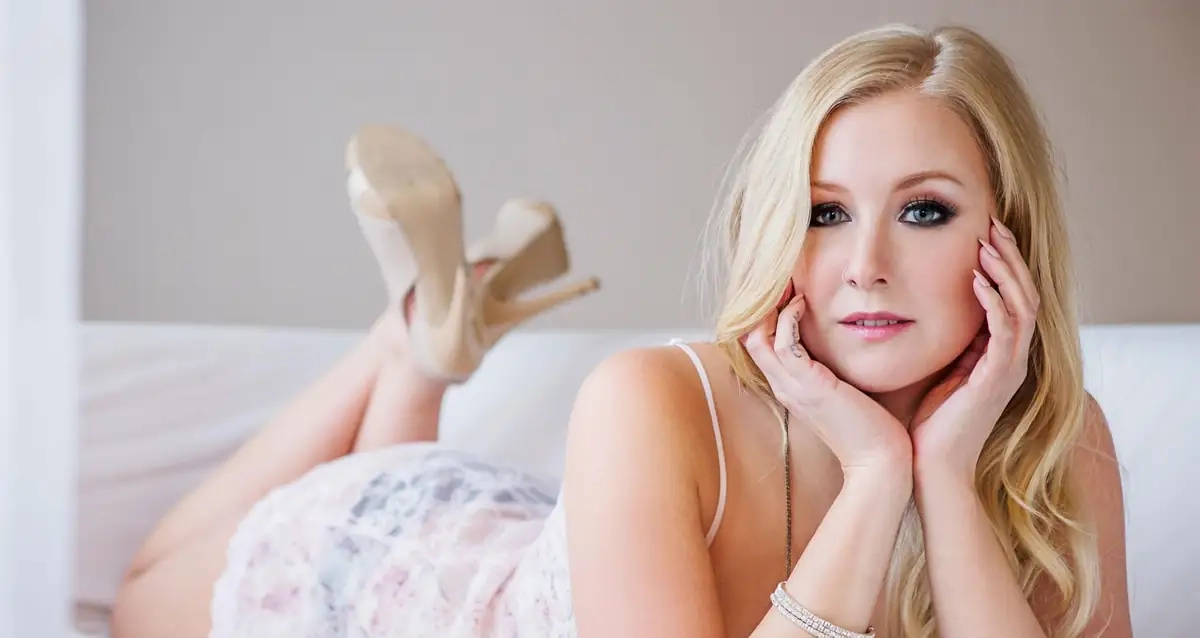 A blonde woman lying on a white sofa in a lace slip, chin in her hands, looking at the camera