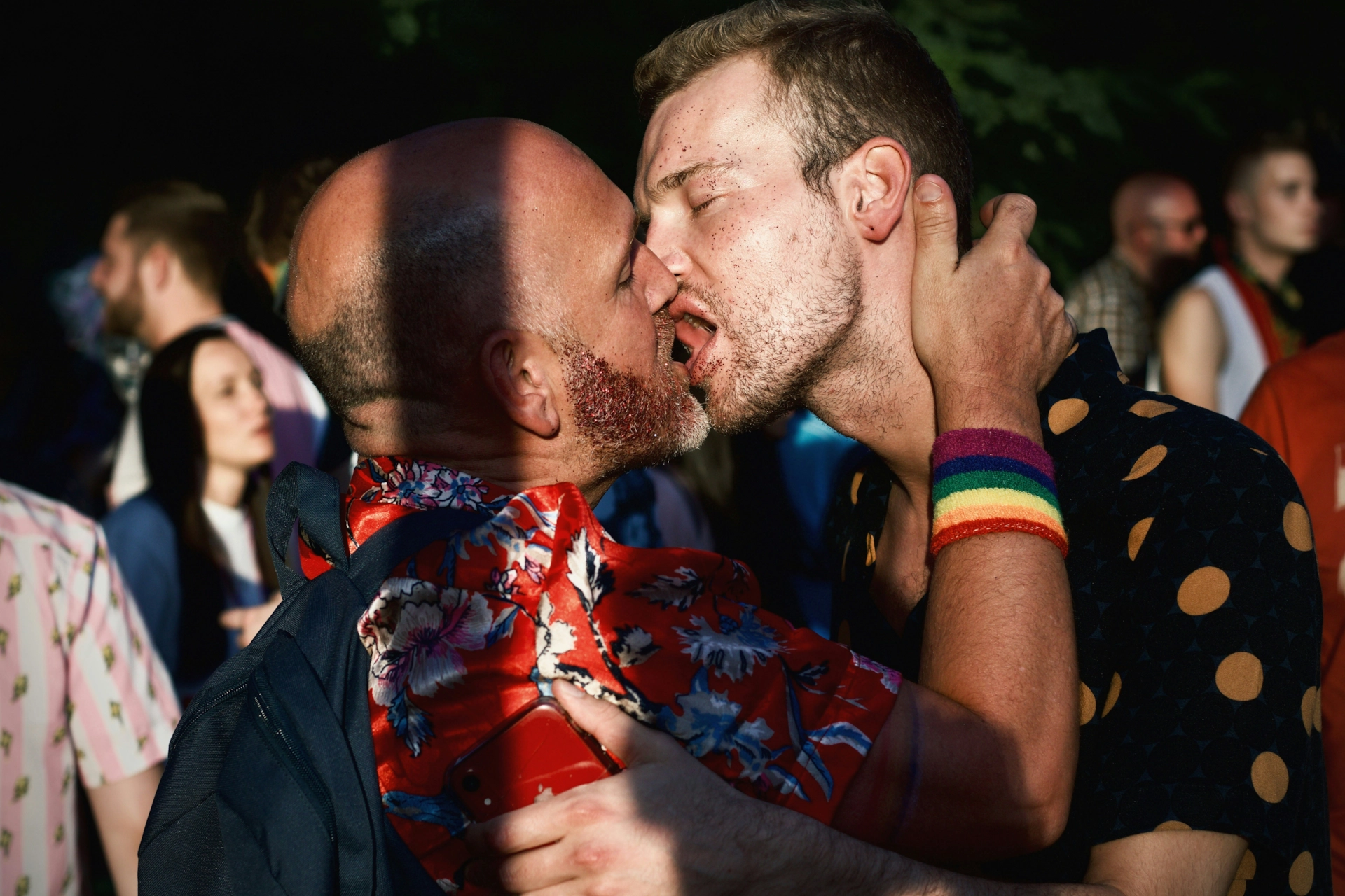Dos hombres besándose en una fiesta, entre la gente.