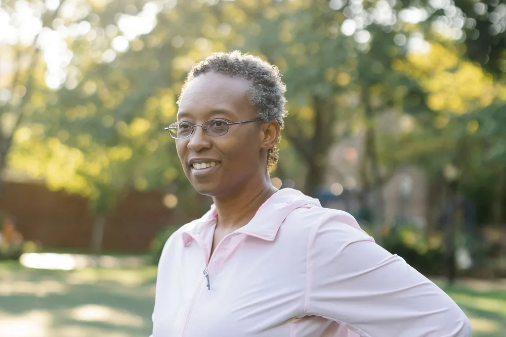 A woman with short grey hair and glasses, in a pale pink jacket, smiling at something off camera in a sunny park