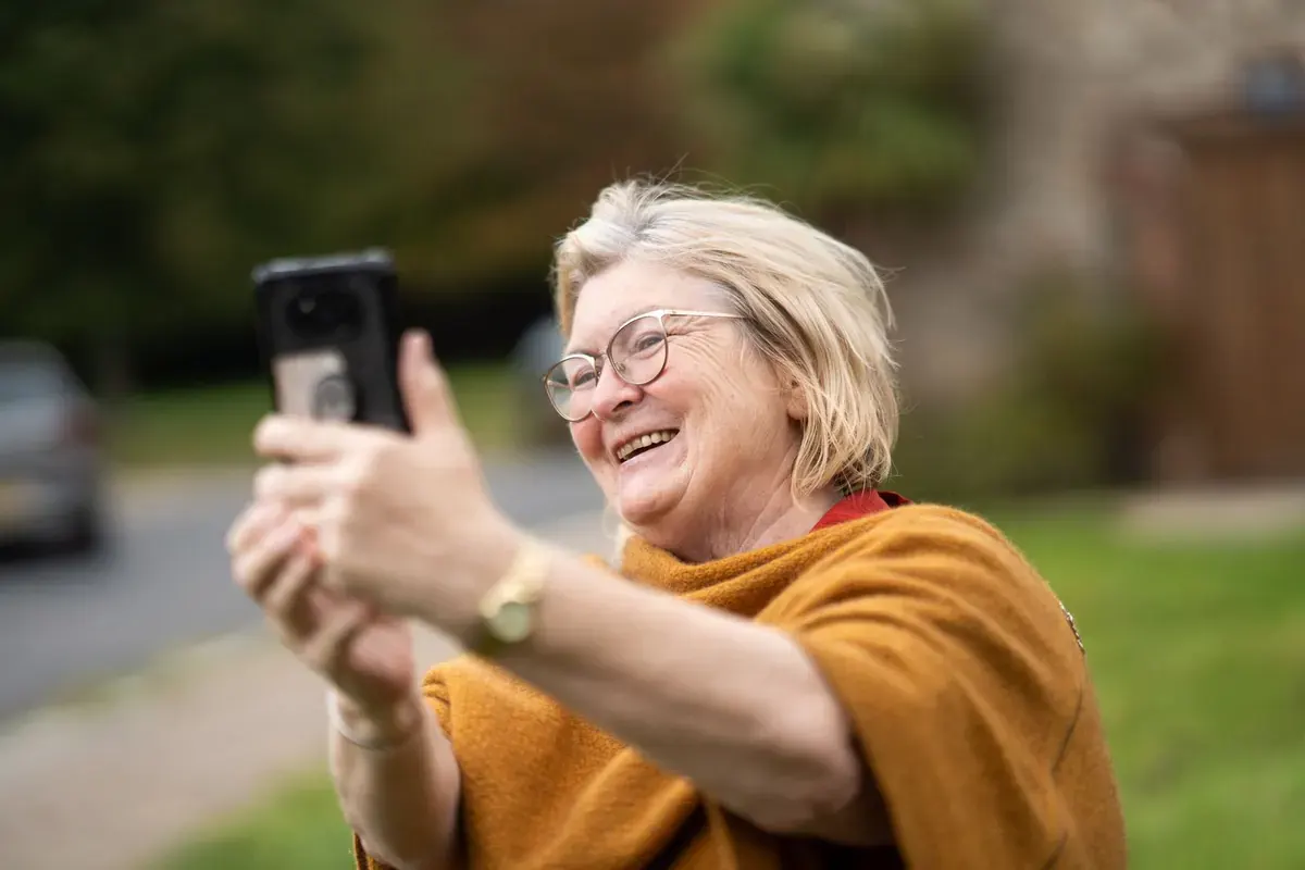 A laughing woman with grey hair and glasses taking a selfie outdoors