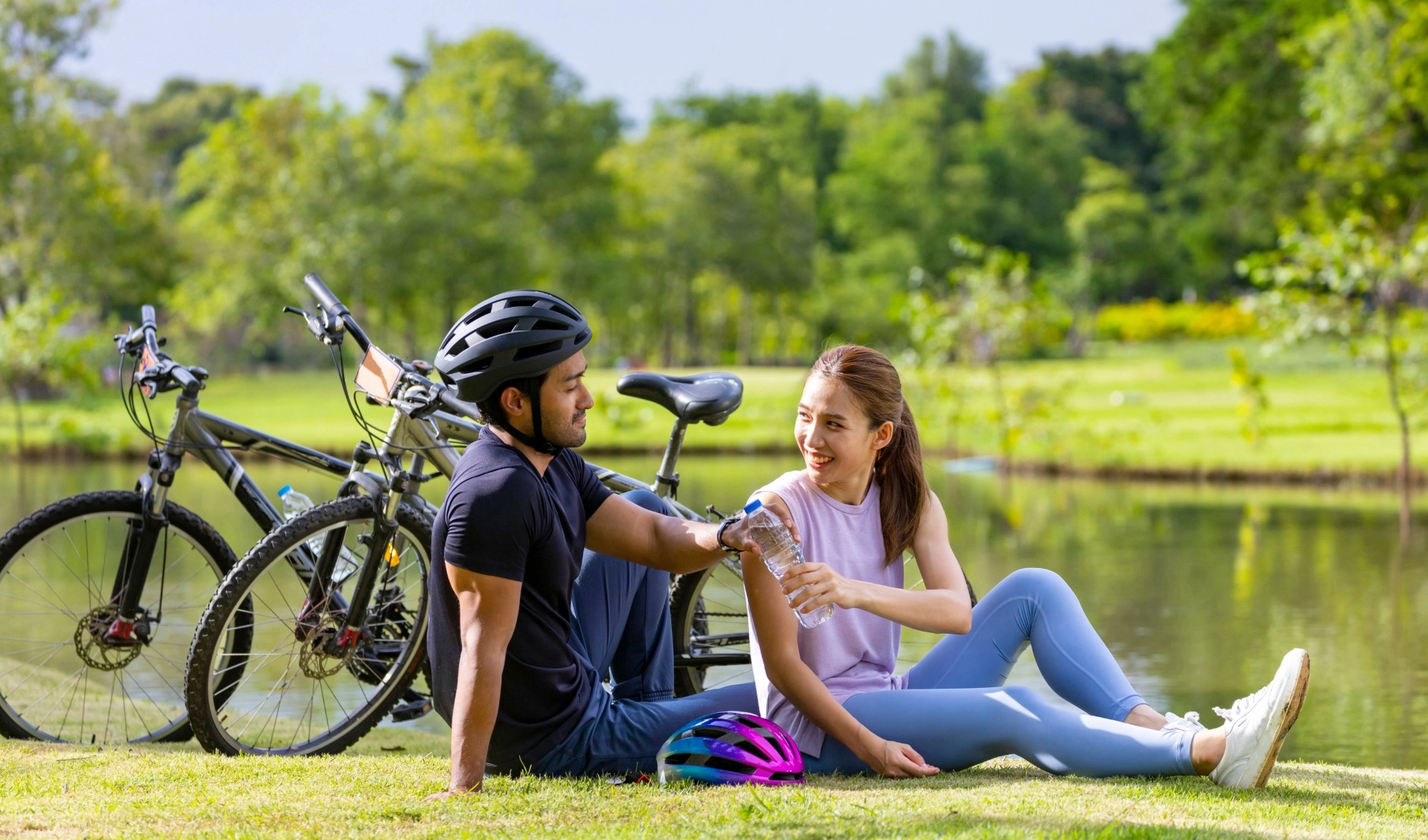 A smiling woman and man taking a break from cycling on the grass beside a lake, with their bikes behind them