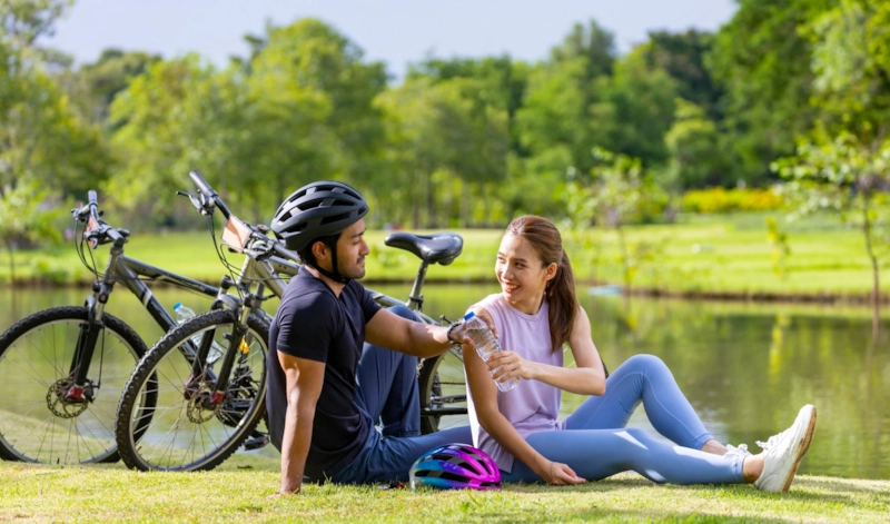 A smiling woman and man resting on the grass by a lake with their bikes