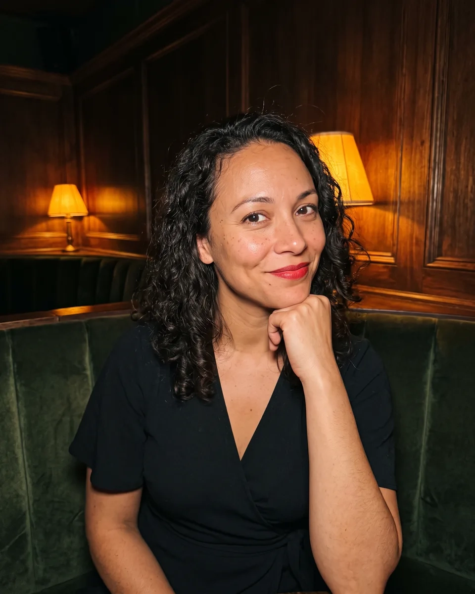A woman with dark shoulder-length curls and red lipstick in a black wrap dress rests her chin on her hand in a green velvet booth of a dimly lit speakeasy-style bar (AI-generated image)
