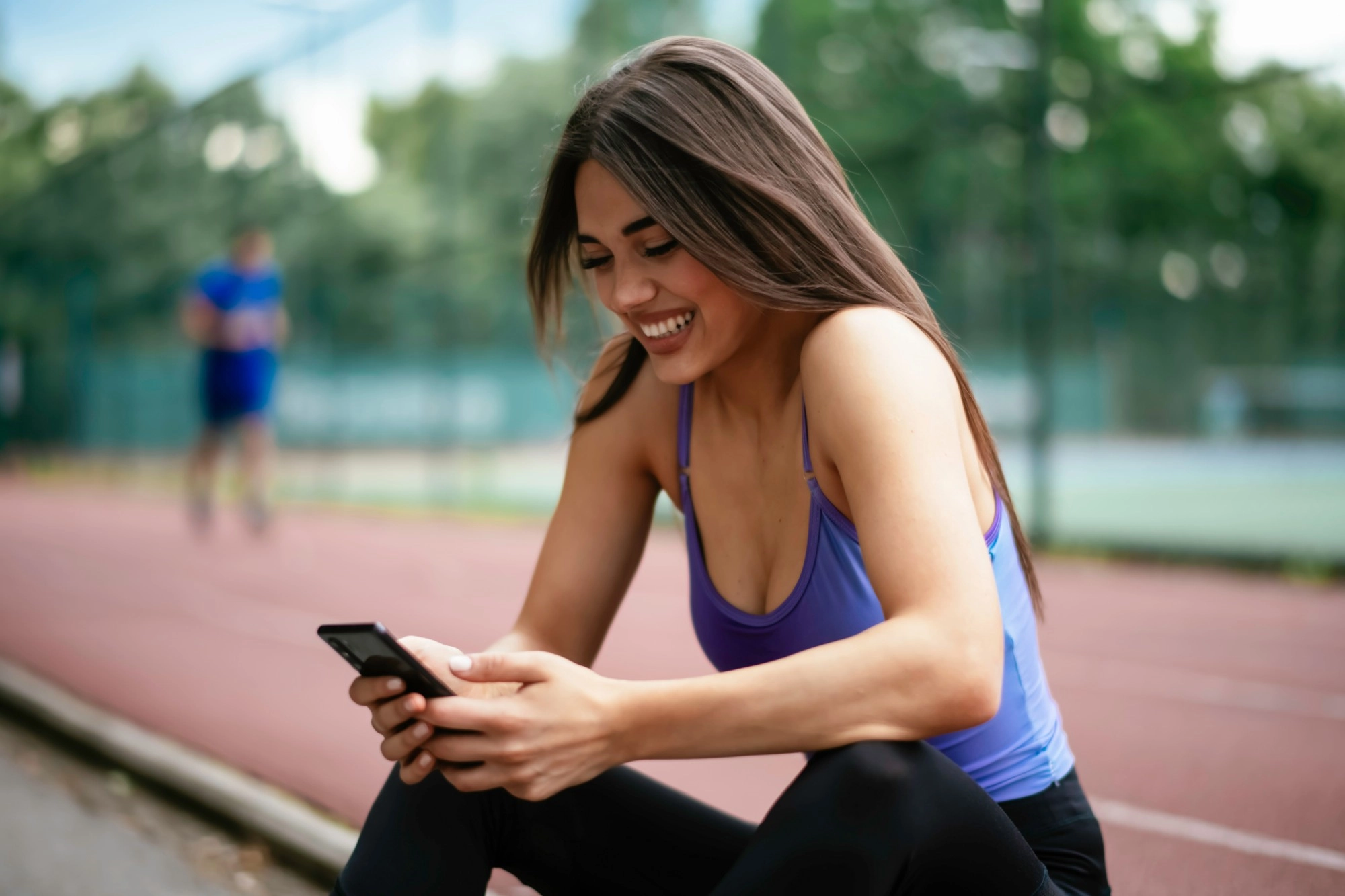 A smiling woman in a purple vest sitting by a running track looking at her phone