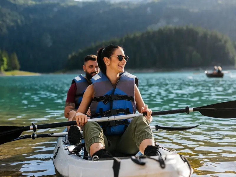 A smiling woman and bearded man in lifejackets paddling a tandem kayak on a turquoise mountain lake