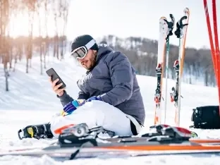 A skier sitting in the snow beside his skis, checking his phone