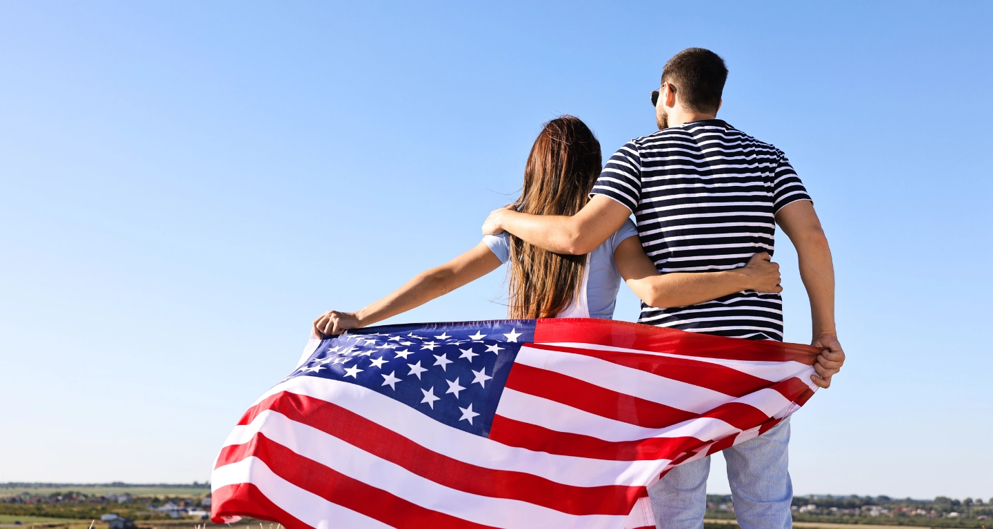 A couple seen from behind with their arms around each other, the American flag wrapped around them under a blue sky