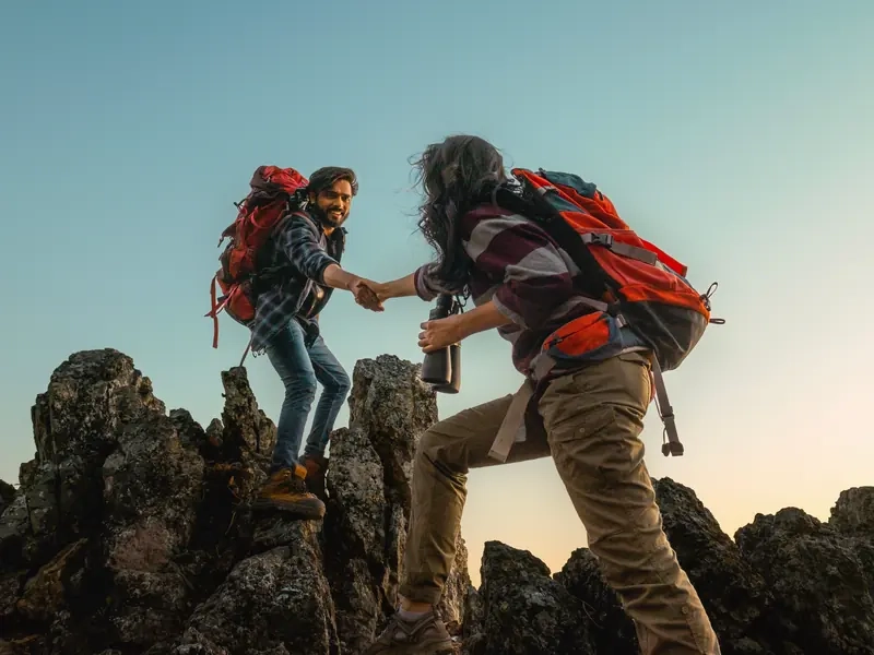 A man and a woman with backpacks clasping hands as they meet on a rocky summit at sunset