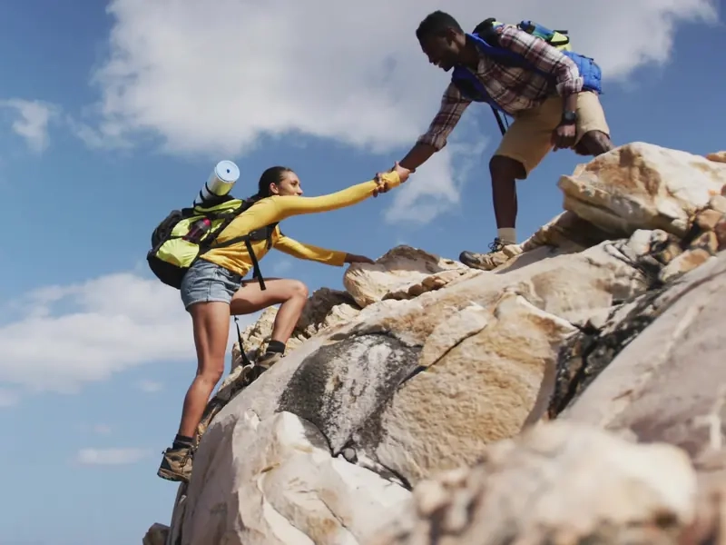 Two hikers with backpacks scrambling up a sunny granite boulder together