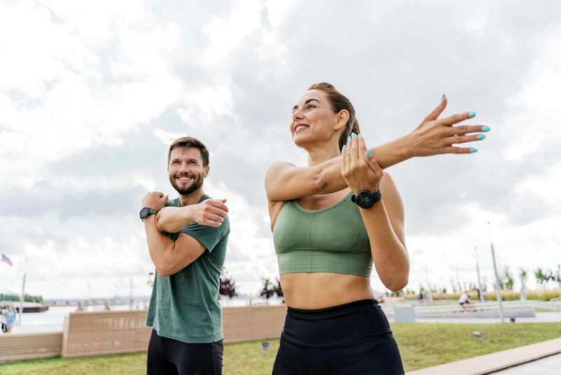 A smiling man and woman stretching their arms together outdoors