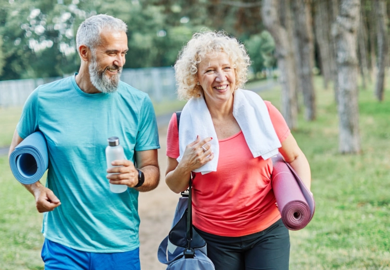 A laughing grey-haired man and curly-haired woman walking in the park with yoga mats