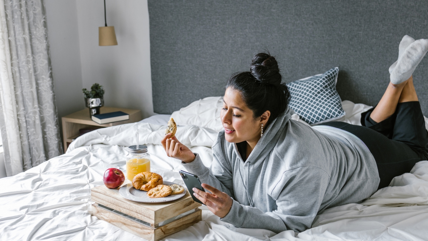 A woman relaxing on her bed with breakfast, checking her phone