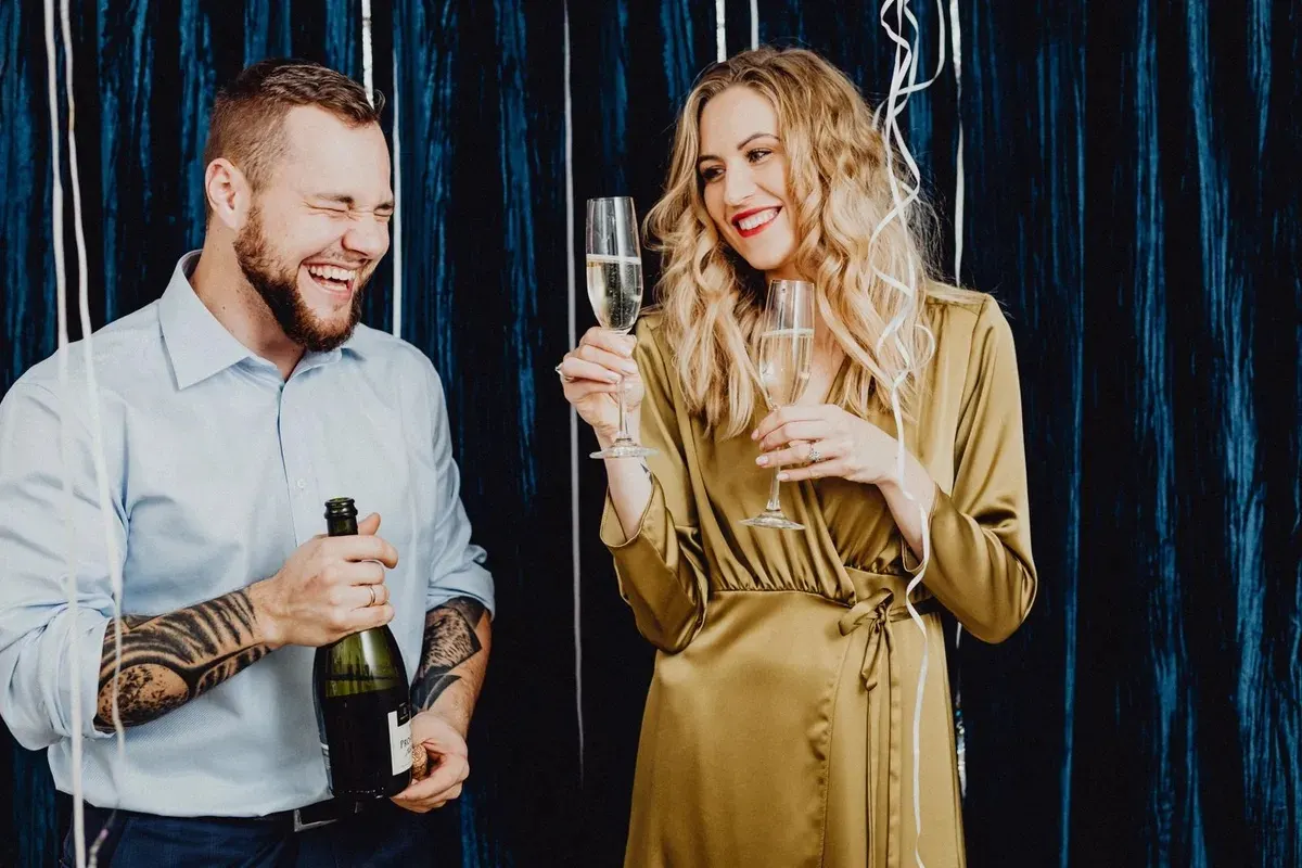 A laughing bearded man holding a champagne bottle beside a blonde woman in a gold satin wrap dress raising her glass at a party