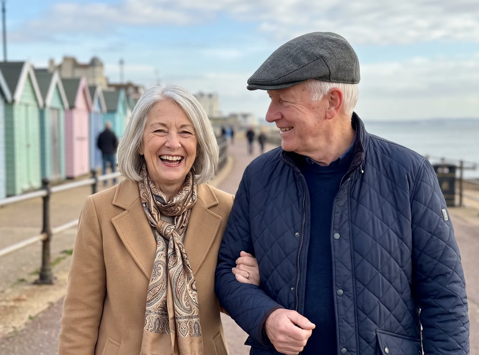 A smiling couple in their sixties walk arm in arm along a seaside promenade lined with pastel beach huts, she in a camel coat, he in a flat cap (AI-generated image)
