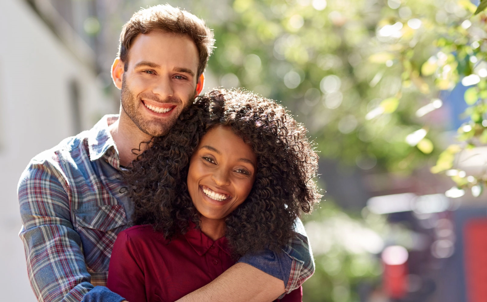 A smiling man with his arms around a smiling woman with curly hair, outdoors
