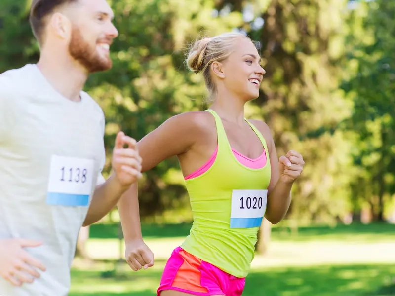 A smiling man and woman wearing race bibs running side by side through a sunny park