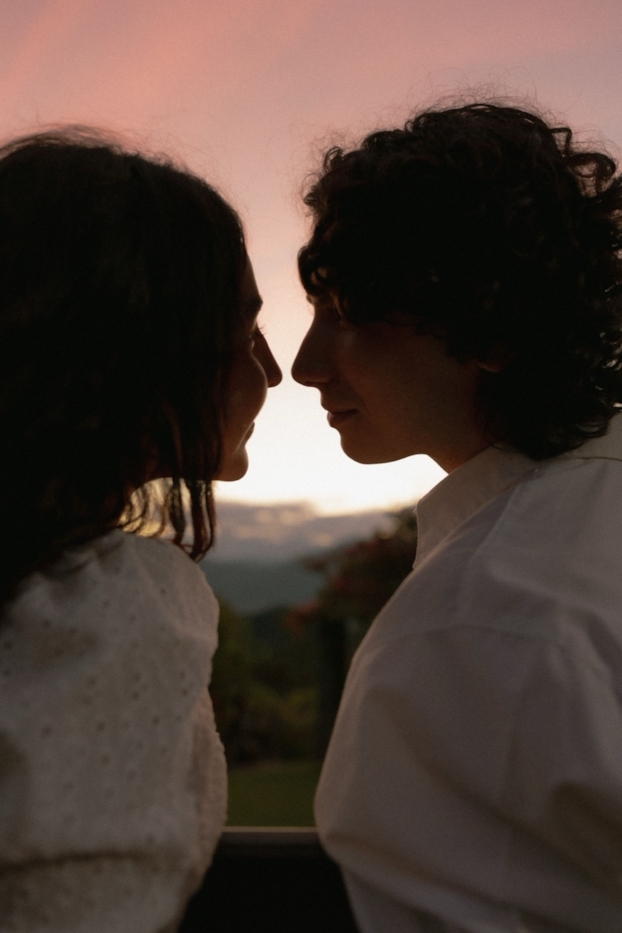 A couple stands in profile at a dusk overlook under warm sunset light