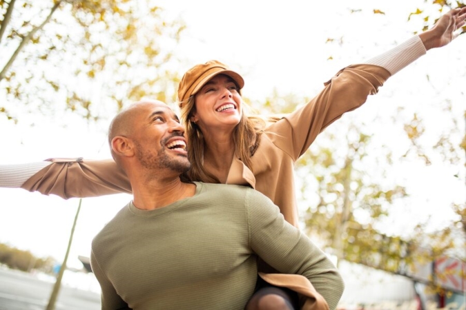 Couple laughing outdoors, woman on man's back with arms spread wide