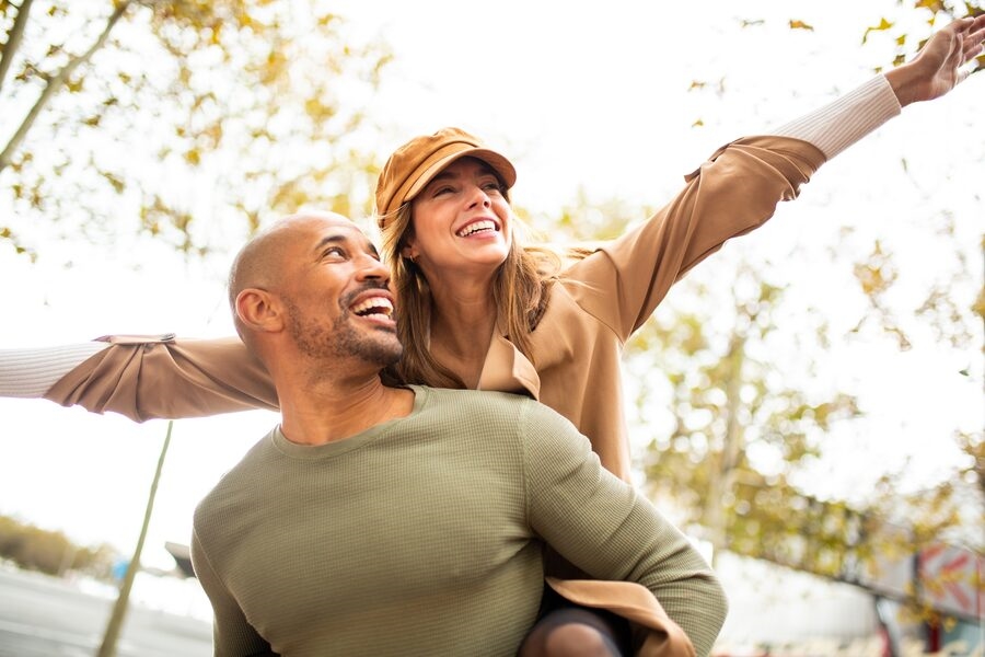 Couple laughing outdoors, woman on man's back with arms spread wide