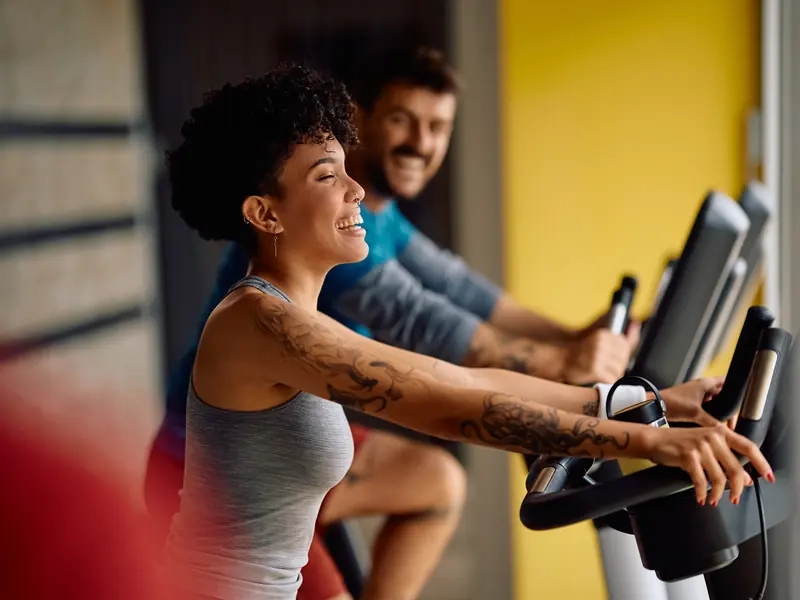 A woman laughing on a spin bike with a smiling man riding behind her