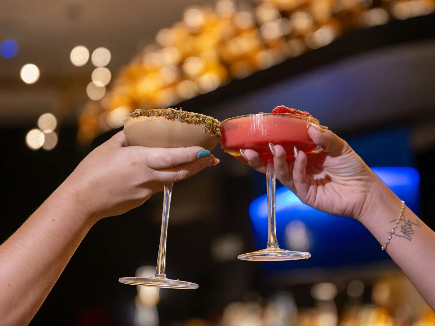 Two hands clinking cocktail glasses in a dimly lit bar, warm lights glowing in the background
