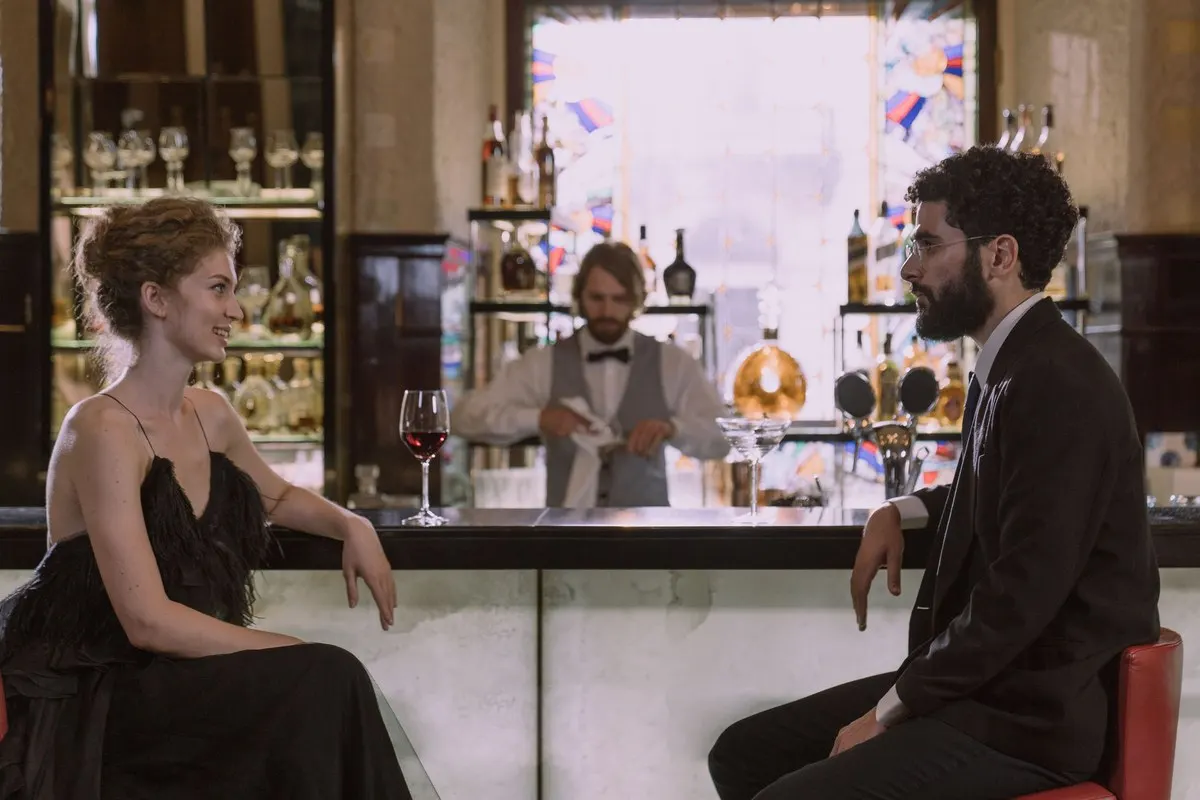 A woman in a black evening dress and a bearded man in a dark suit sitting at a hotel bar, smiling at each other while the bartender works behind them