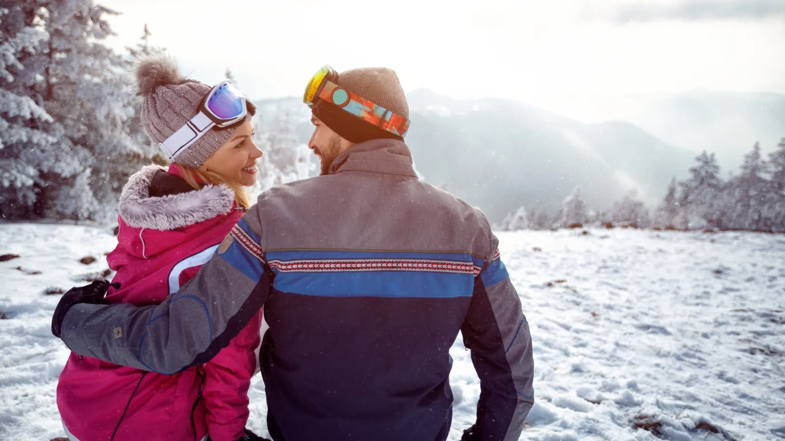 A couple in knit hats and goggles, arm in arm, smiling at each other on a snowy mountain lookout