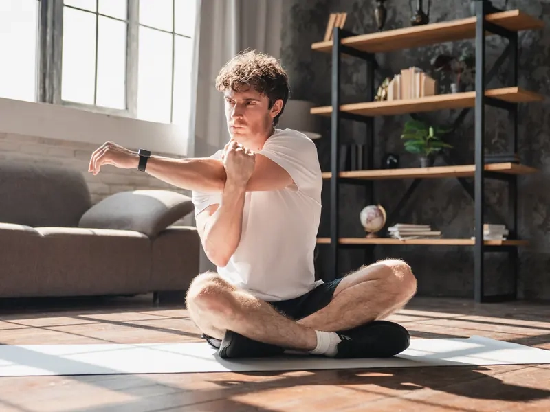 A man sitting cross-legged on a yoga mat at home, stretching his arm in morning light