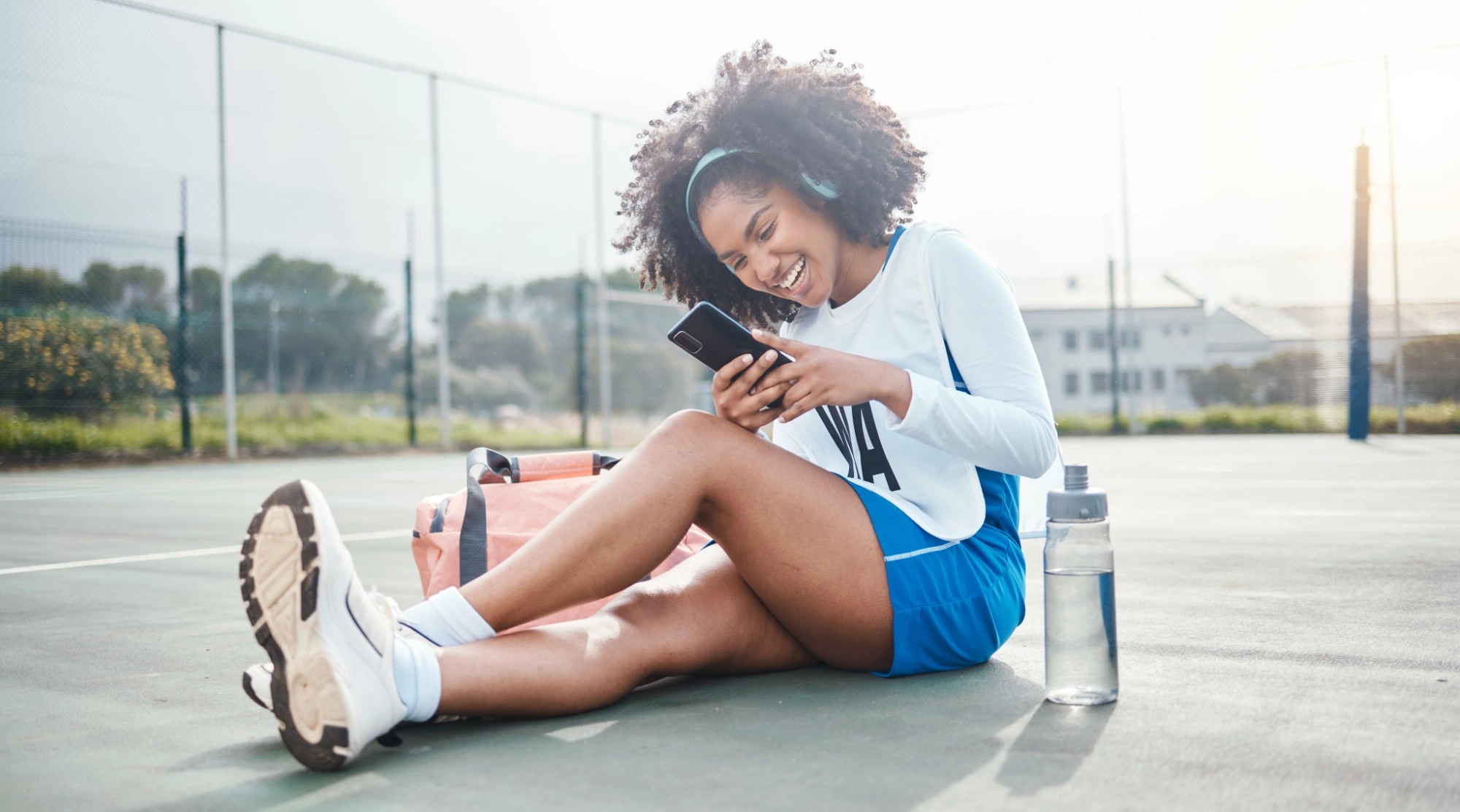 A laughing woman in headphones sitting on a netball court looking at her phone