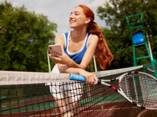 A smiling woman leaning on a tennis net, holding her racket and phone