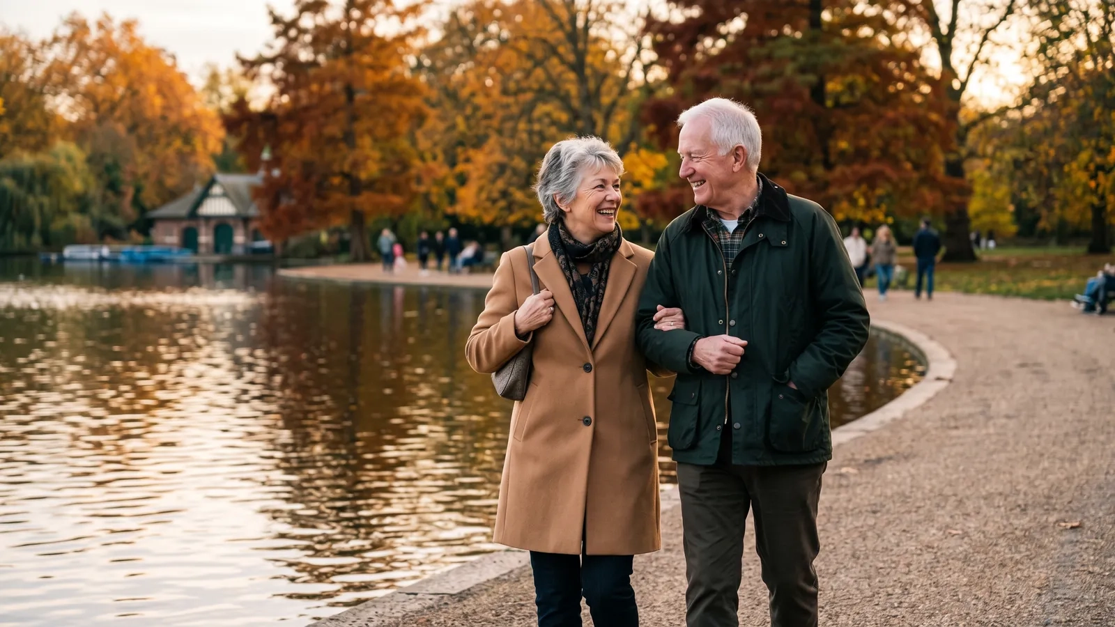 A couple in their late sixties walk arm in arm along a lakeside path in a large park with autumn trees, smiling at each other (AI-generated image)