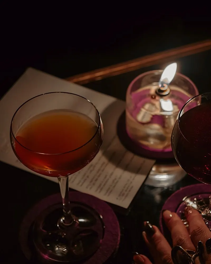 A cocktail glass and a flickering candle on a dark bar table, a printed menu beside them and a hand resting at the edge of the frame