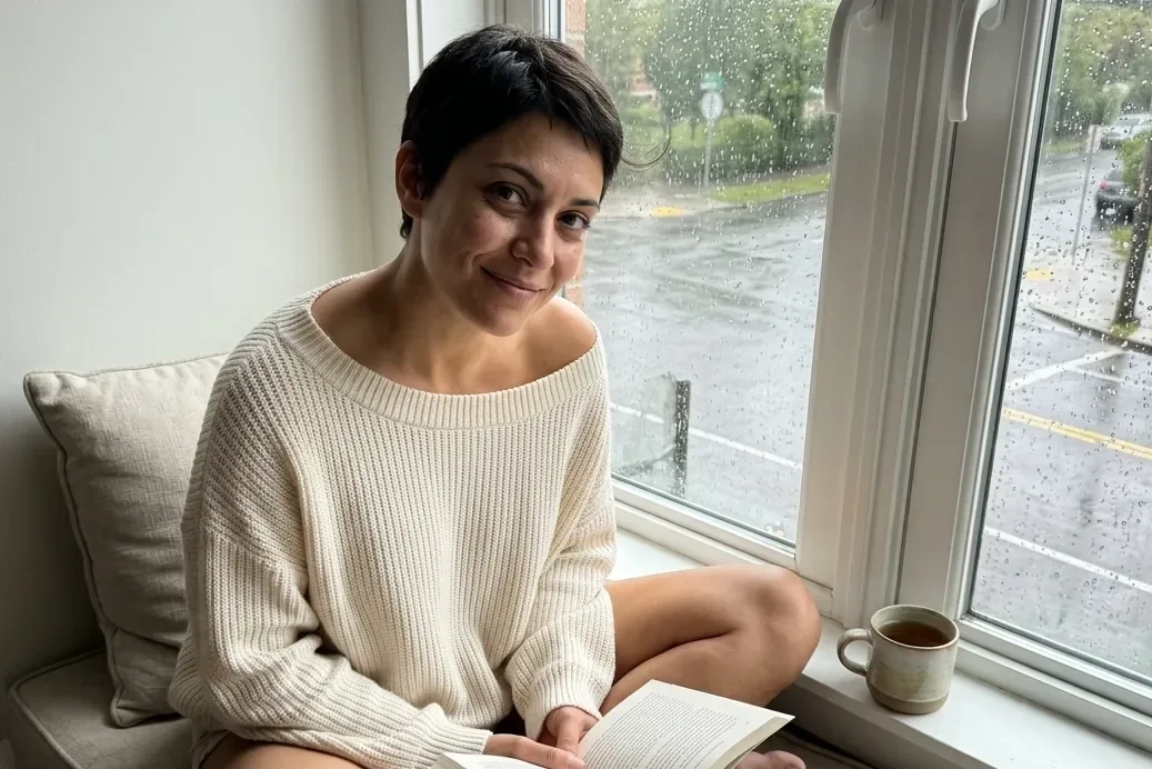 A woman with a short dark pixie cut and an off-the-shoulder cream sweater sits cross-legged on a window seat with an open book and a mug of tea, smiling at the camera while rain runs down the window (AI-generated image)