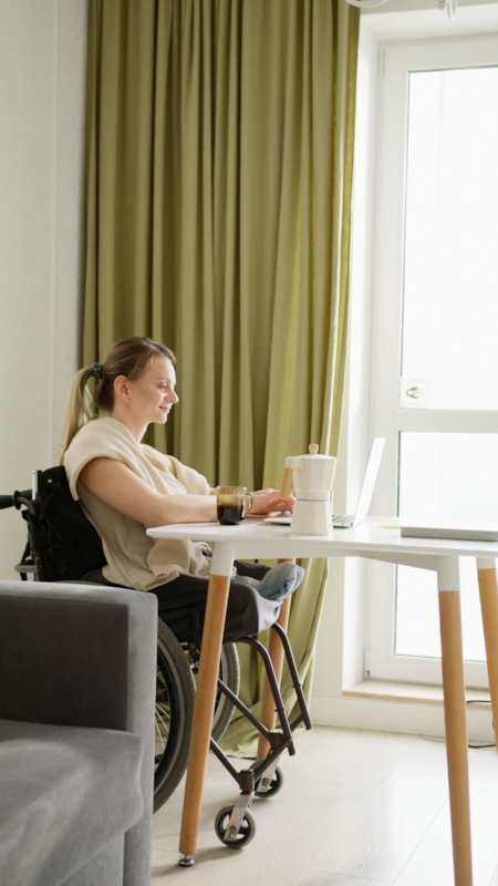 A woman in a wheelchair smiling while typing on a laptop at a table by a sunlit window
