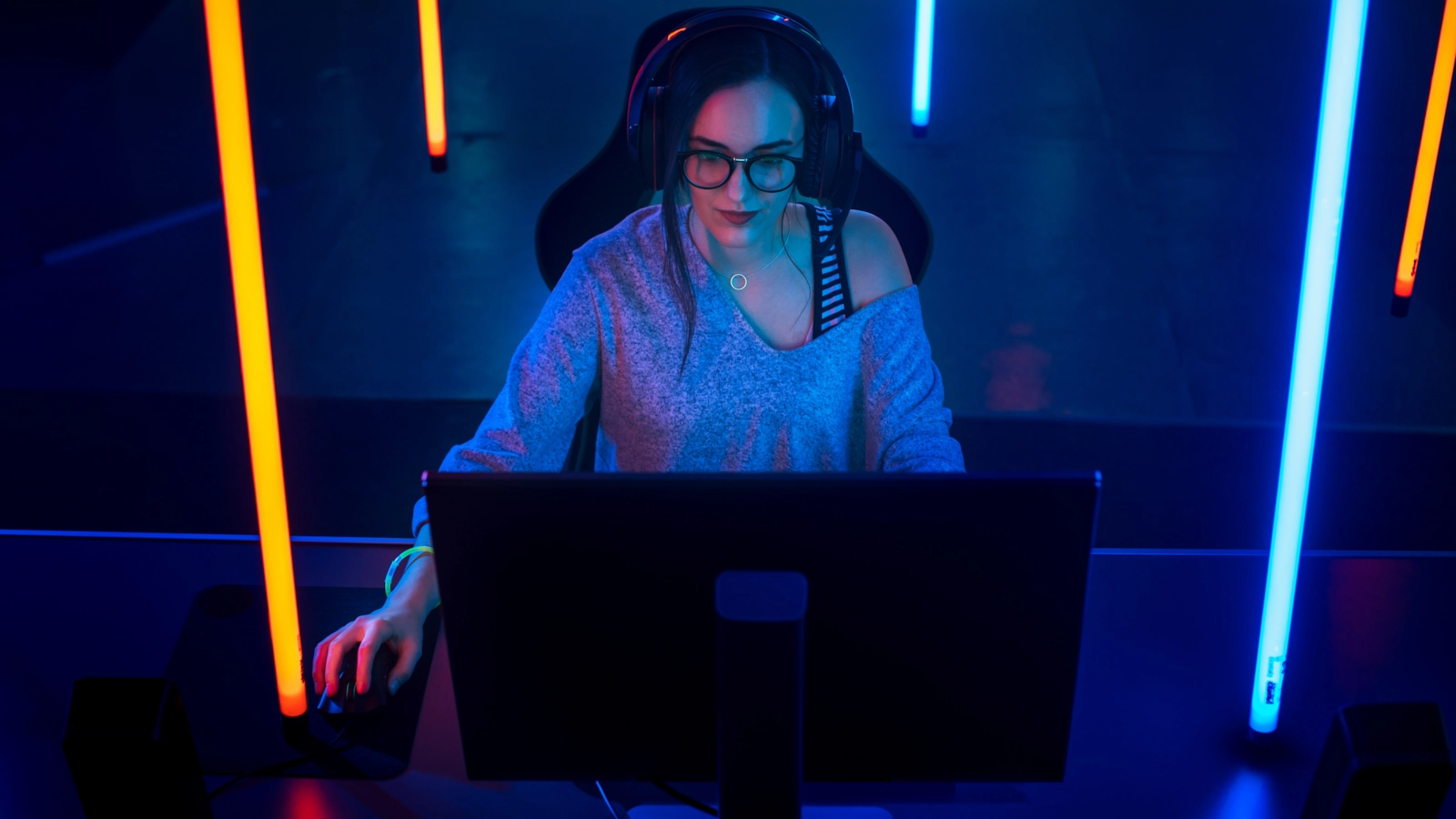 A woman in glasses and a headset gaming at a monitor in blue and orange neon light