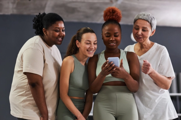 Four smiling women of different ages in workout clothes looking at a phone together in a studio