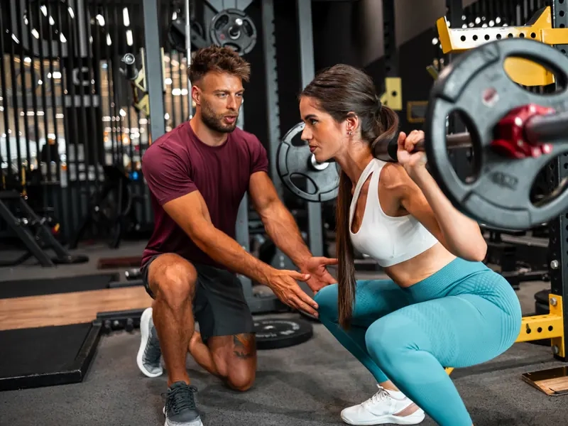 A trainer coaching a woman through a barbell back squat in a gym
